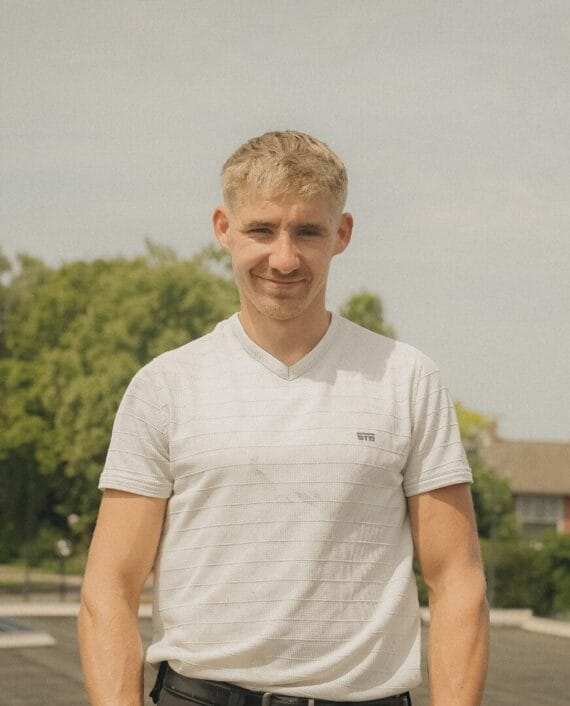 Een jonge man met kort blond haar staat buiten en draagt een wit gestreept t-shirt, met bomen en een gebouw op de achtergrond.