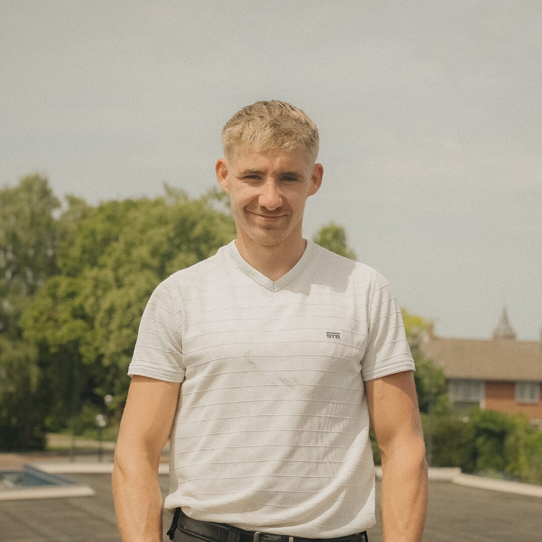 Een jonge man met kort blond haar staat buiten en draagt een wit gestreept t-shirt, met bomen en een gebouw op de achtergrond.
