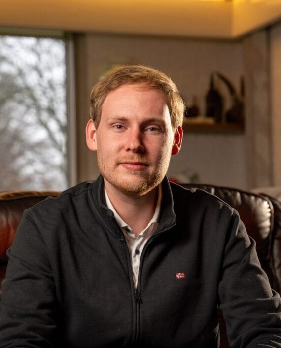 Een man met een korte baard zit op een leren bank, draagt een donker jack met rits over een overhemd met kraag. Op de achtergrond zijn een raam en een plank met decoratie-items te zien.