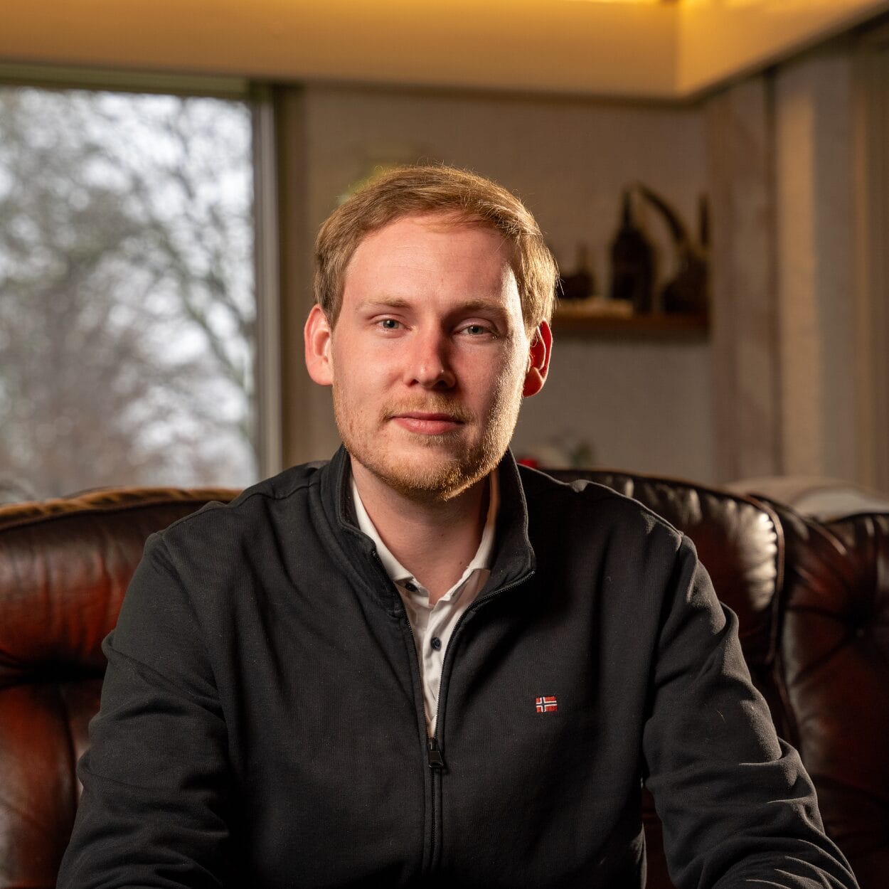 Een man met een korte baard zit op een leren bank, draagt een donker jack met rits over een overhemd met kraag. Op de achtergrond zijn een raam en een plank met decoratie-items te zien.
