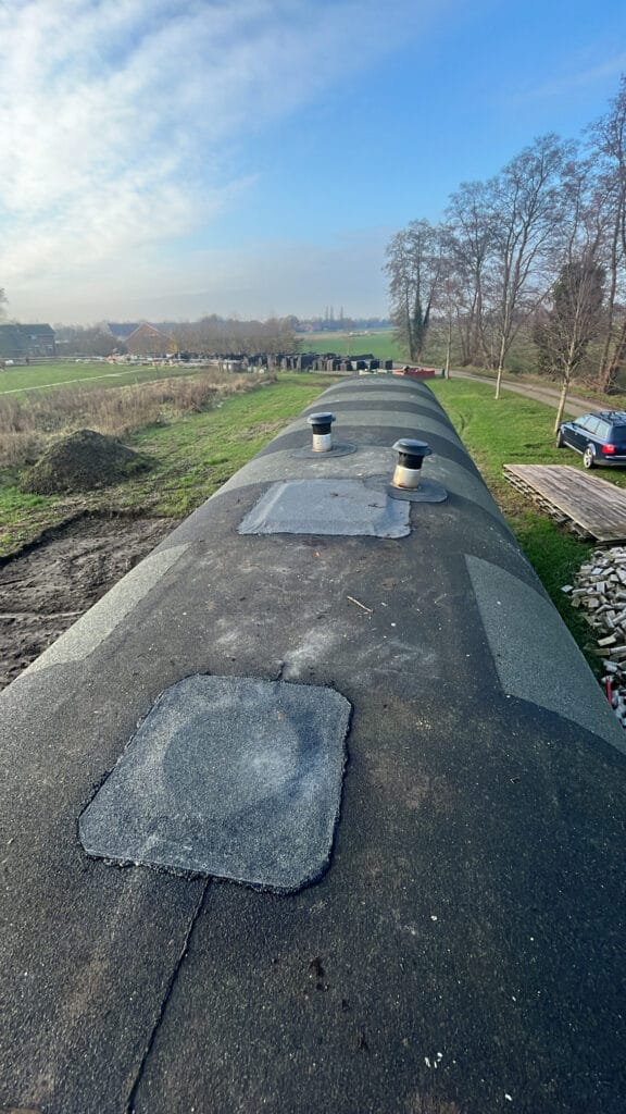 Een geteerde, bunkerachtige constructie met ventilatieopeningen, omgeven door een landelijk landschap; een auto op de achtergrond, met velden en een stapel hout in de buurt.