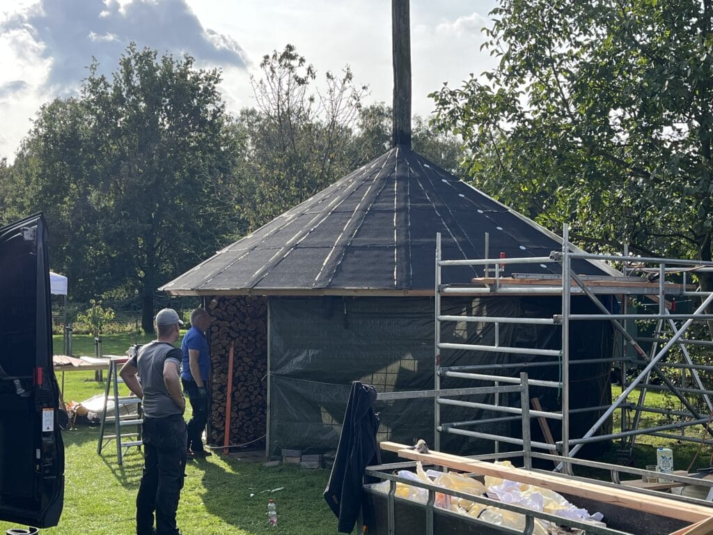 Twee arbeiders staan bij een gedeeltelijk gebouwd rond gebouw met een schuin dak en steigers in een grasveld. Er staan boomstammen tegen het gebouw gestapeld.