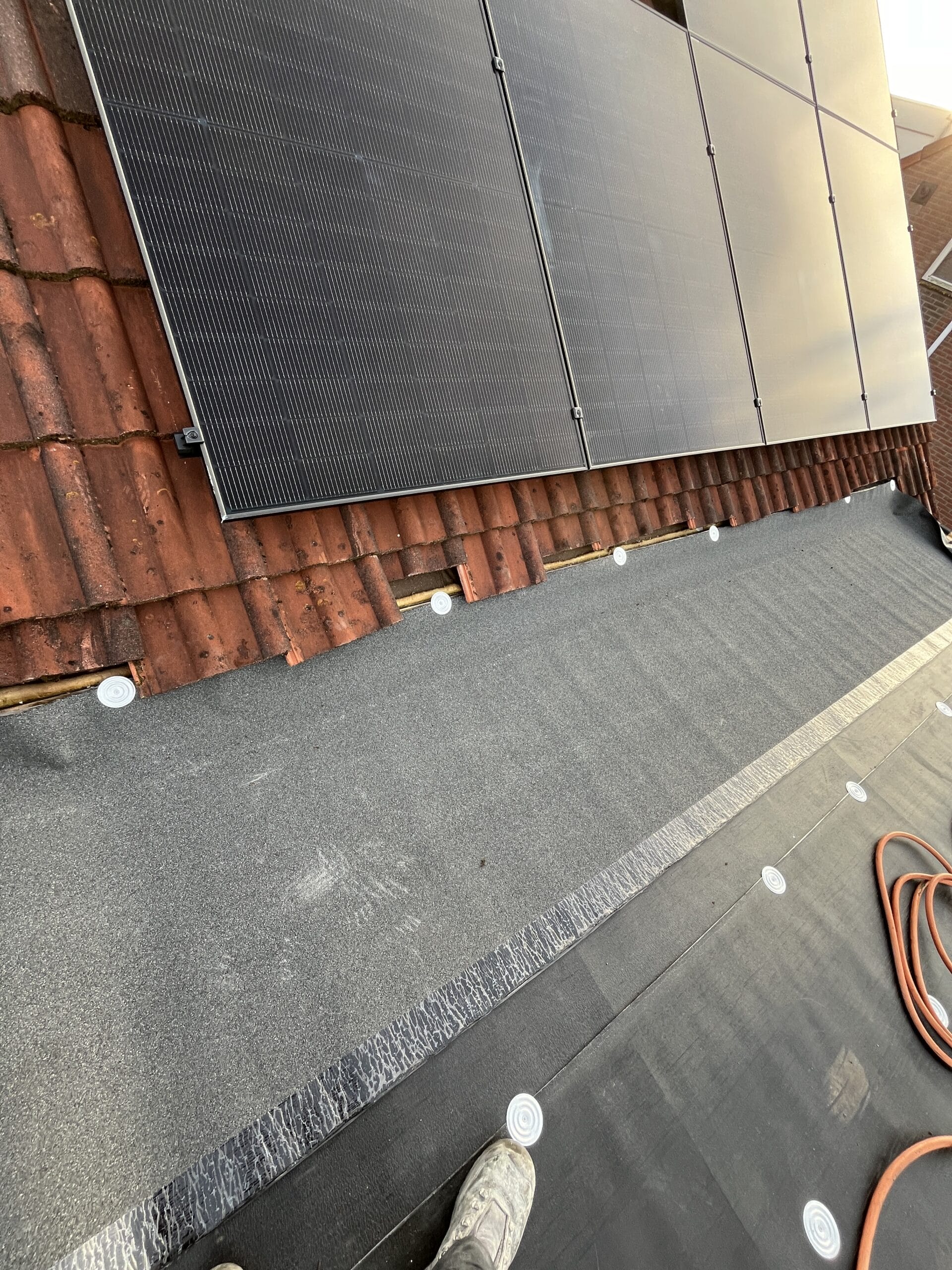 Dakaanzicht met geïnstalleerde zonnepanelen en dakbedekkingsmaterialen, waaronder teerpapier en dakpannen. Een voet gedeeltelijk zichtbaar op de voorgrond.