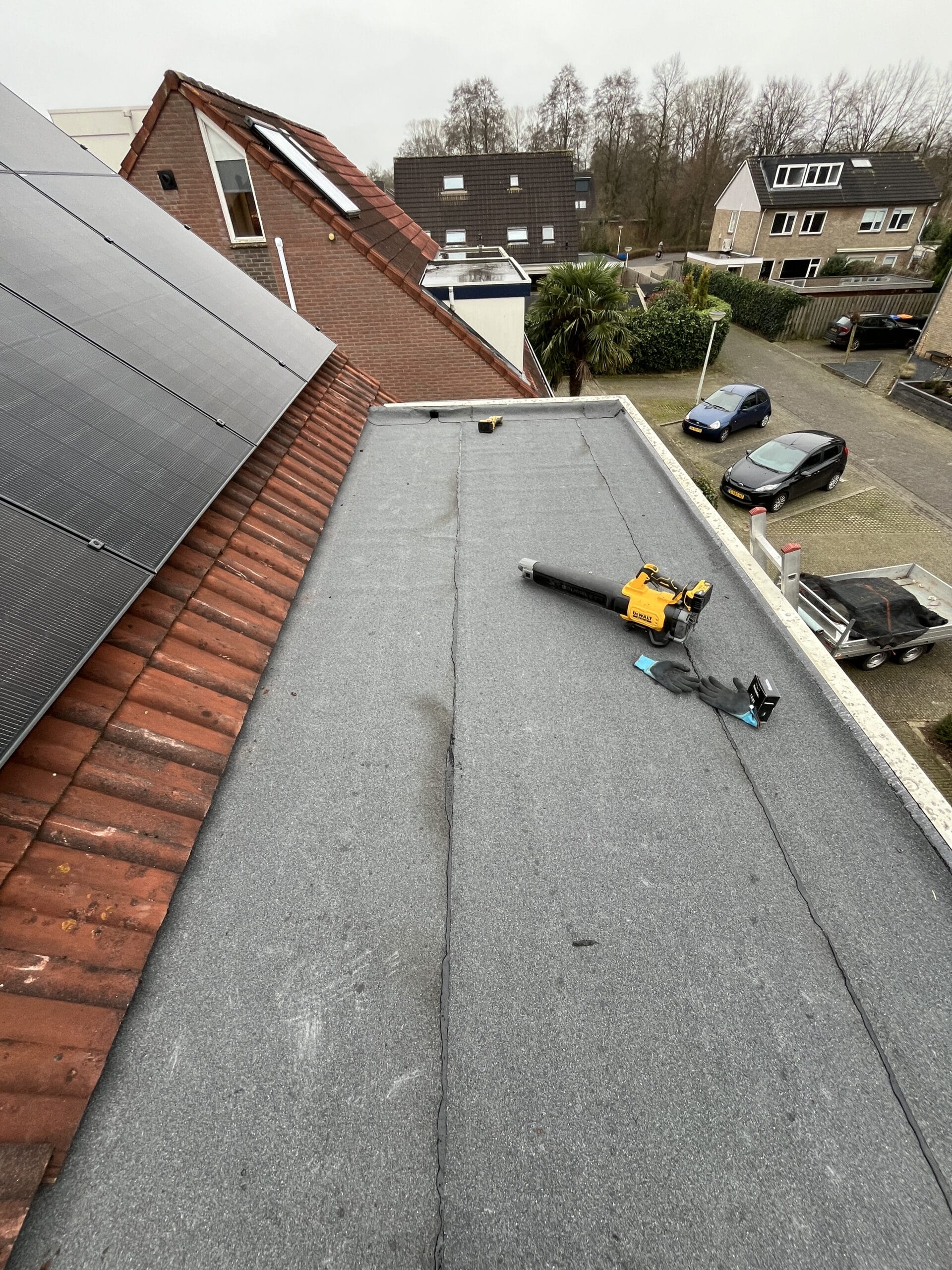 Plat dak met grijze bedekking, een bladblazer en dakgereedschap. Huizen met zonnepanelen zijn zichtbaar, naast geparkeerde auto's en bomen op de voorstedelijke achtergrond.