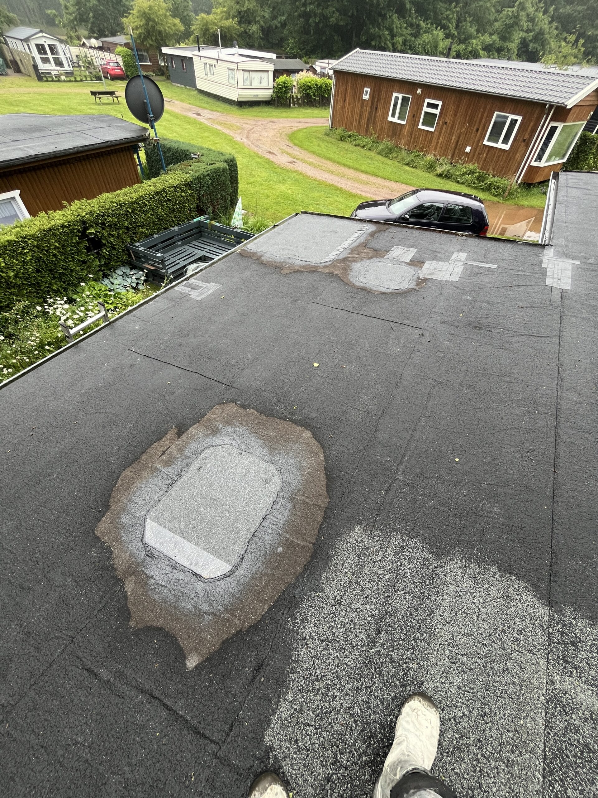 Uitzicht op een plat dak met reparaties, omgeven door groen en houten gebouwen. Een geparkeerde auto is zichtbaar bij de constructie.