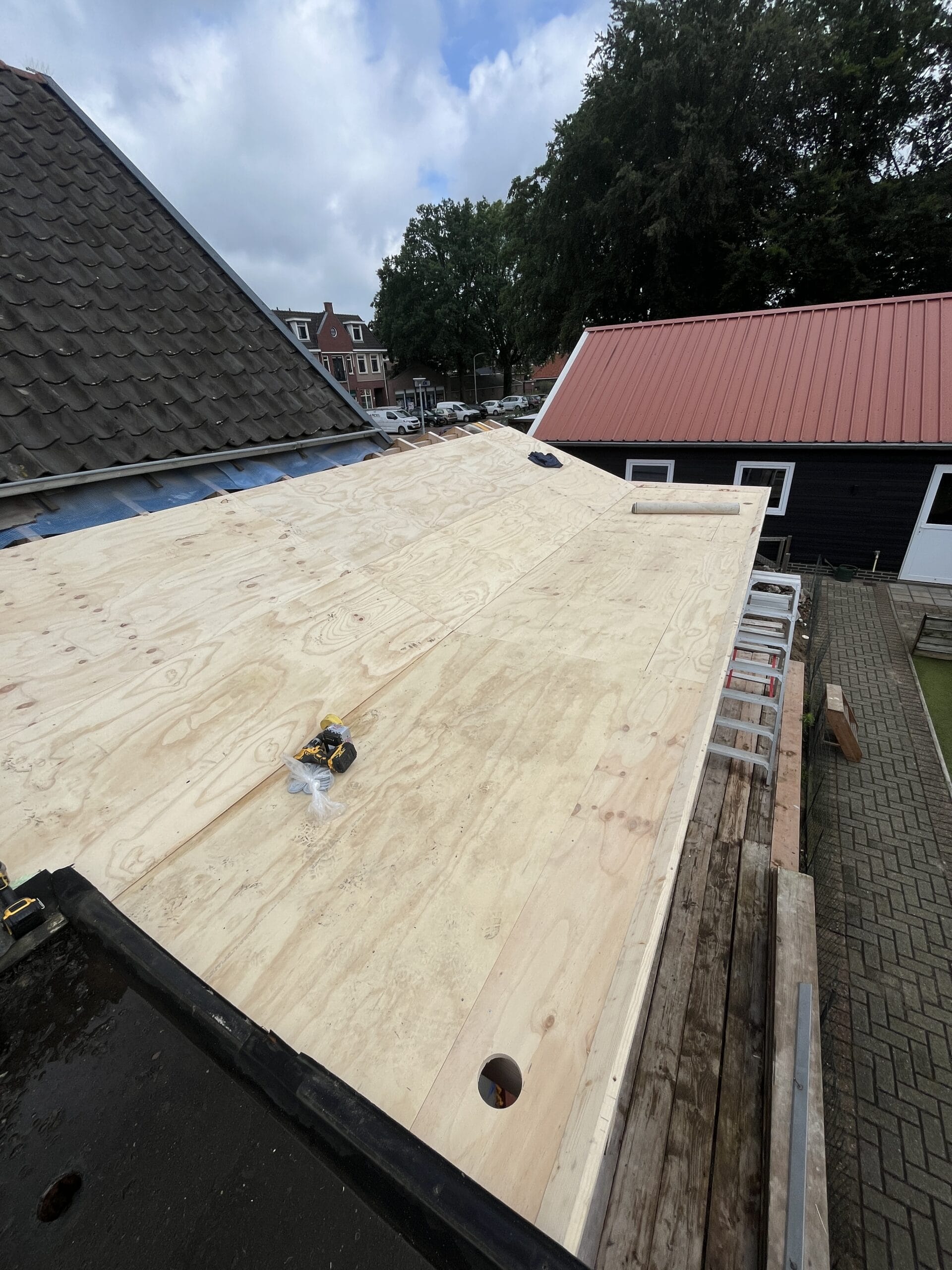 Onder een bewolkte lucht zijn multiplexplaten op een dakframe gemonteerd, met zichtbaar gereedschap en twee aangrenzende gebouwen op de achtergrond.