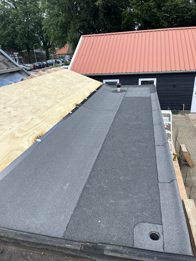 Bouwterrein met de installatie van een plat dak met grijze asfaltgordels, naast een gebouw met een rood dak.
