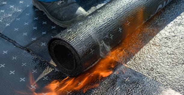 Een rol dakisolatie wordt in brand gestoken op een plat dak, met zichtbare vlammen eronder. Er staat een persoon met een laars in de buurt.