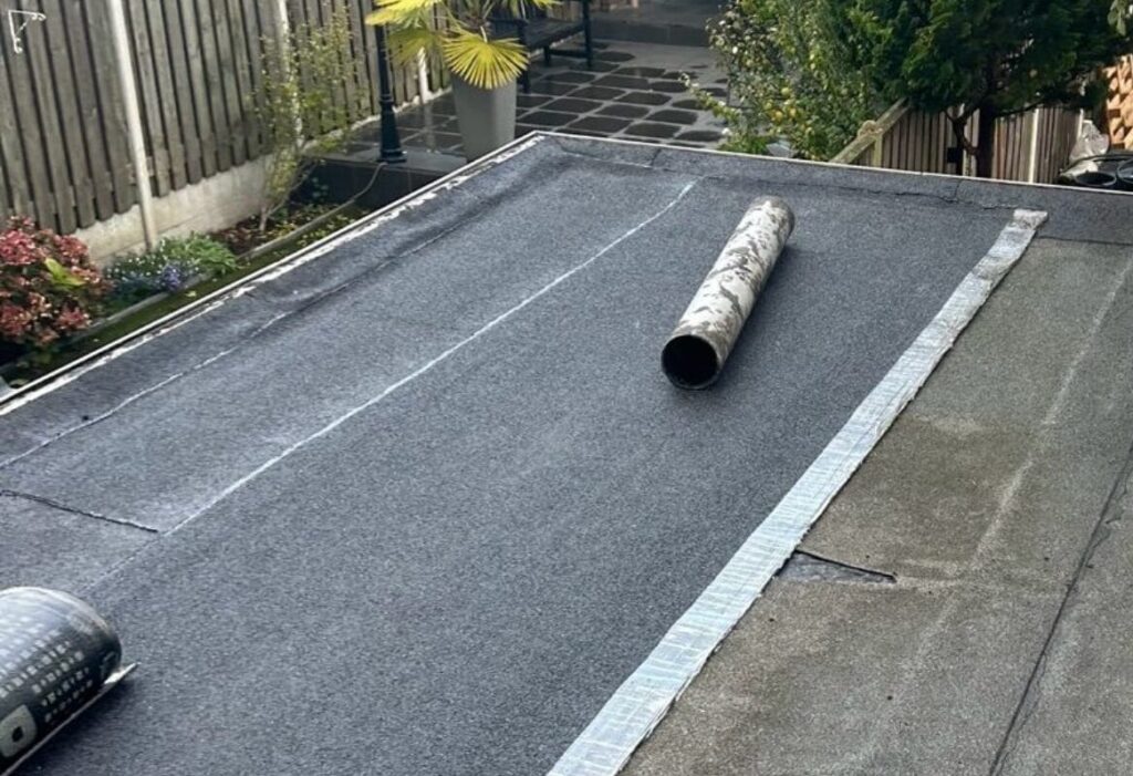 Plat dak met een gedeeltelijk afgerolde rol dakbedekking, op de achtergrond zijn de tuin en het hekwerk zichtbaar.