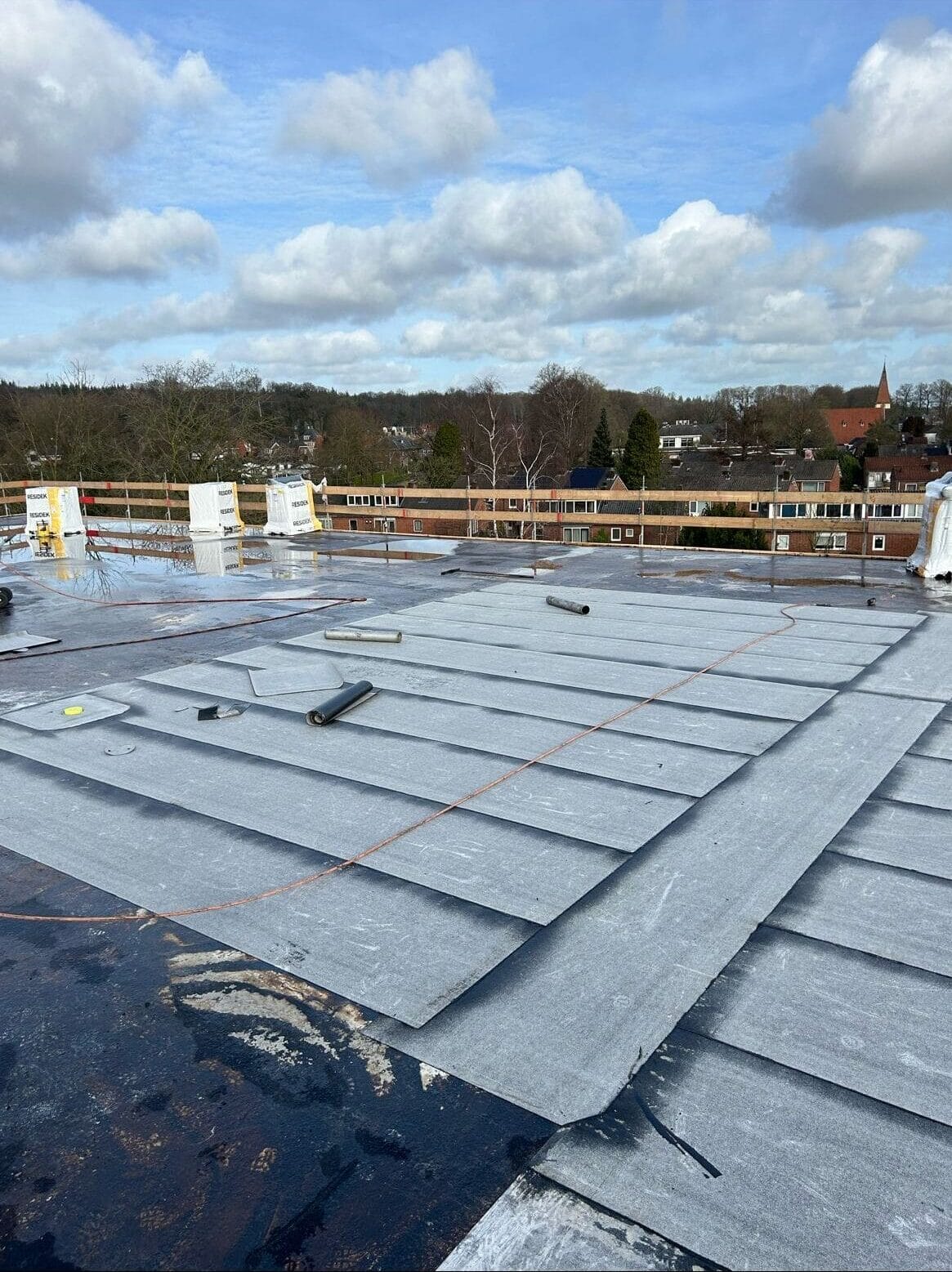 Dak met een gedeeltelijk geïnstalleerd waterdicht membraan, verspreid gereedschap en uitzicht op een bewolkte lucht en verre gebouwen.