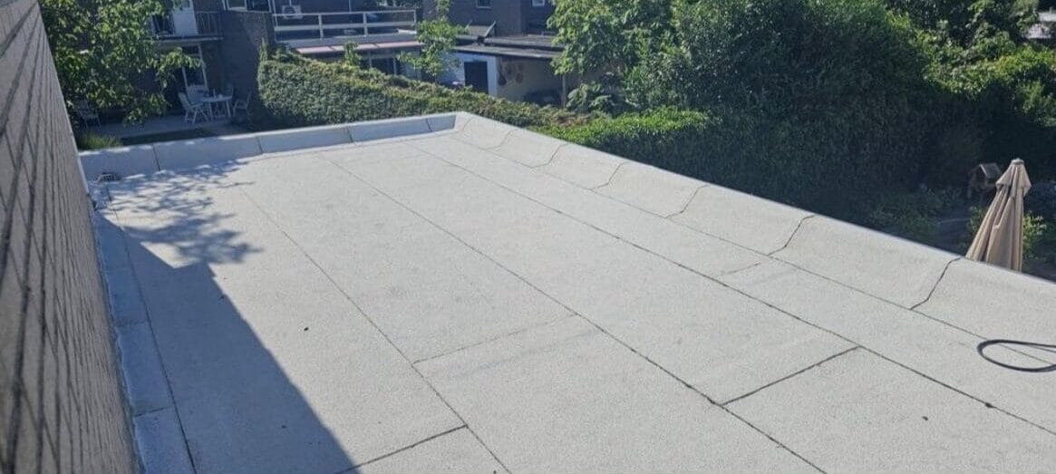 Dakinspectie voor plat dak met betonnen oppervlak, begrensd door een bakstenen muur aan de linkerkant. Bomen en gebouwen zijn zichtbaar op de achtergrond onder een heldere hemel.