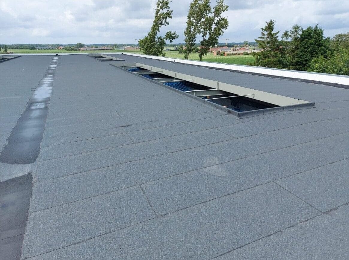 Plat grijs dak met rechthoekige dakramen, perfect voor daklekkage opsporen. Bomen en velden zijn zichtbaar op de achtergrond onder een bewolkte lucht.