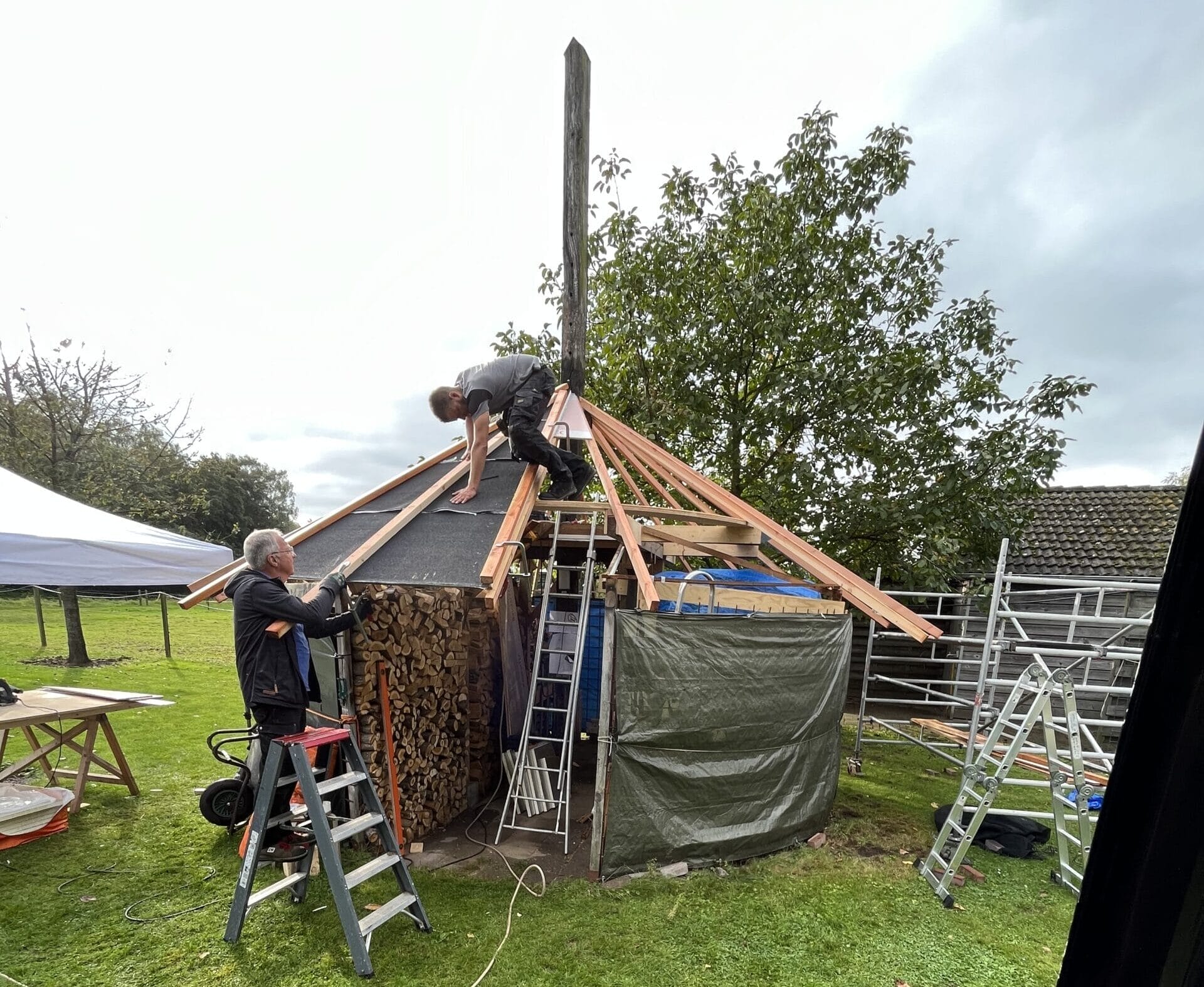 Twee mensen werken nauwgezet aan de constructie van een kleine, ronde houten constructie. Eén persoon staat op een ladder, terwijl de ander balanceert op het dakframe, waarvan een deel onder een zeil ligt. Het is bijna alsof ze een geïmproviseerde dakinspectie uitvoeren met steigers in de buurt ter ondersteuning.