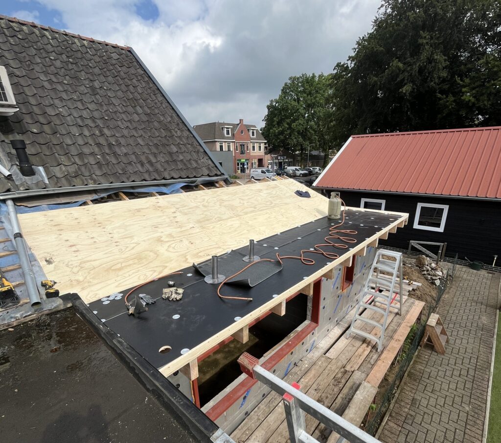 Een dak in aanbouw met multiplex bekleding en gereedschap verspreid over de dakbedekking, onderdeel van een lopende dakrenovatie, naast een schuin pannendak en steigers. Op de achtergrond zijn huizen en bomen zichtbaar.