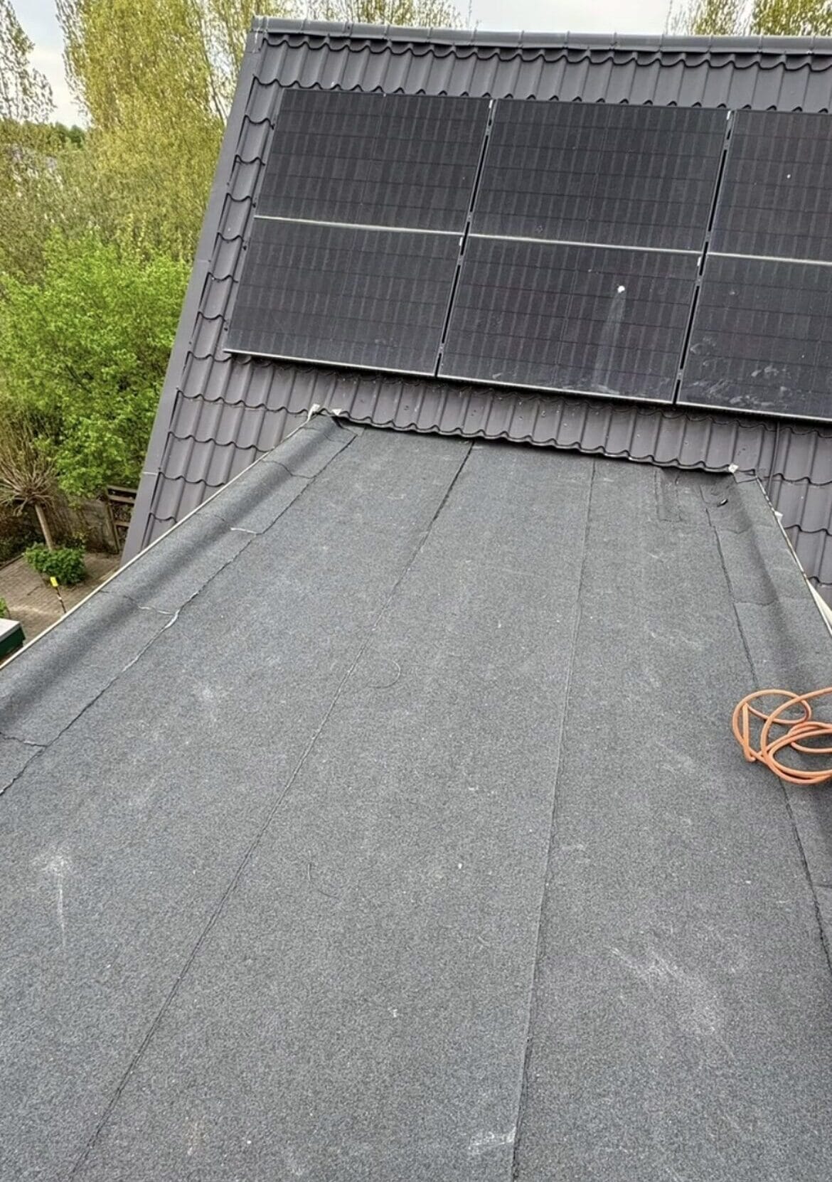 Een schuin dak bedekt met donker dakbedekking, een oranje verlengsnoer en zonnepanelen op het bovenste gedeelte duiden erop dat het dak recent is onderhouden. Op de achtergrond zijn bomen en struiken zichtbaar.