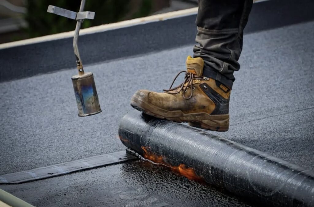 Een persoon met werkschoenen staat op de dakbedekking terwijl een brander de rand verhit en deze met precisie op het dakoppervlak afdicht.