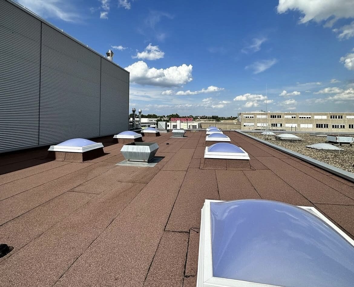 Plat dak met verschillende rechthoekige dakramen, met Roofiva design, grenzend aan een groot gebouw onder een blauwe hemel met verspreide wolken.