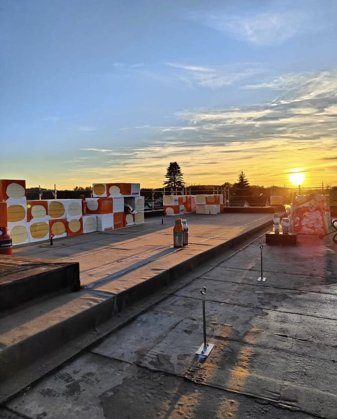 Zonsondergang boven een bouwplaats met gestapelde isolatiematerialen en betonnen oppervlakken, werpt lange schaduwen op het dak tijdens een recente dakinspectie.