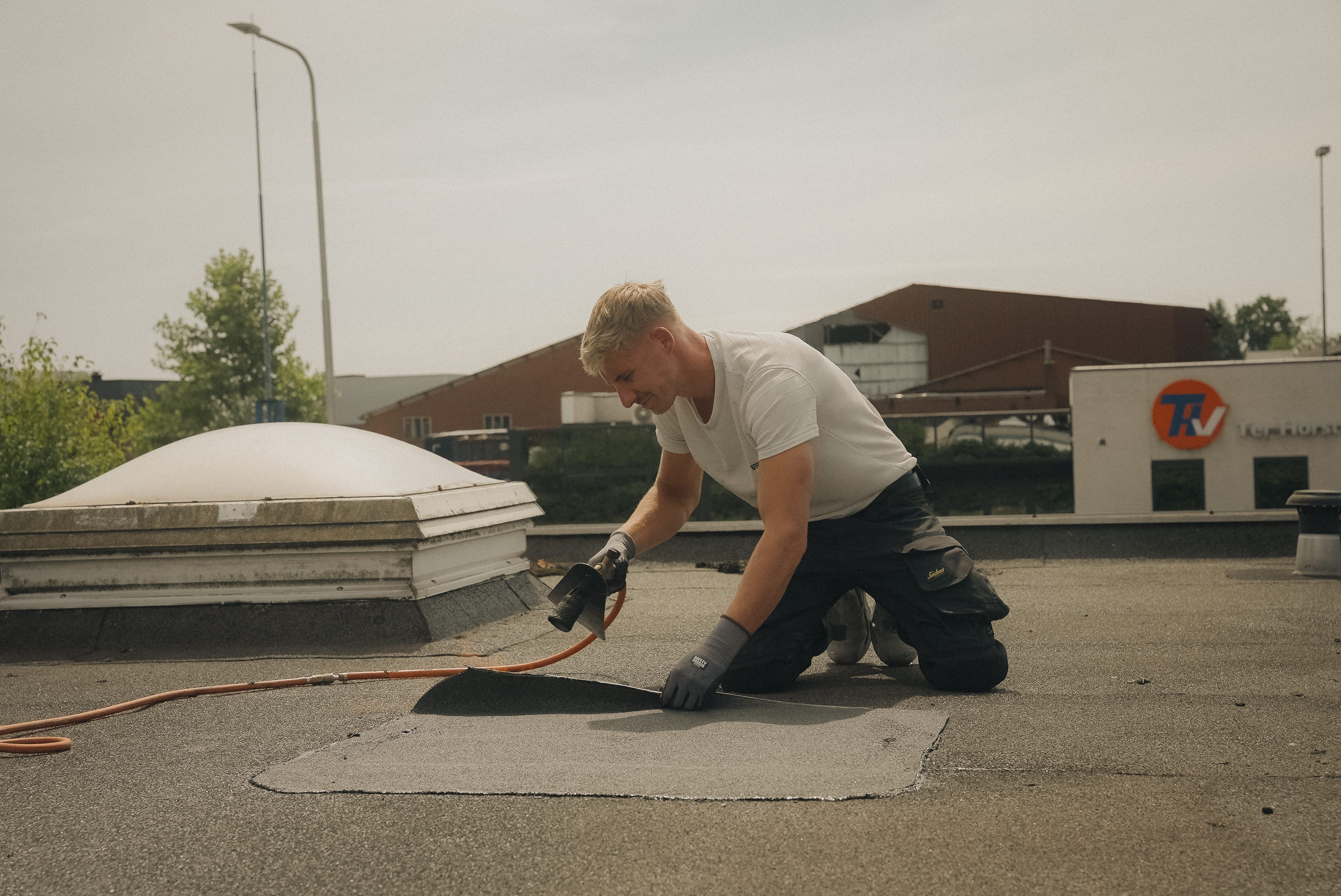 Een arbeider in een wit shirt en werkbroek gebruikt een gereedschap om dakbedekking aan te brengen of af te dichten op een plat dak onder een heldere hemel.