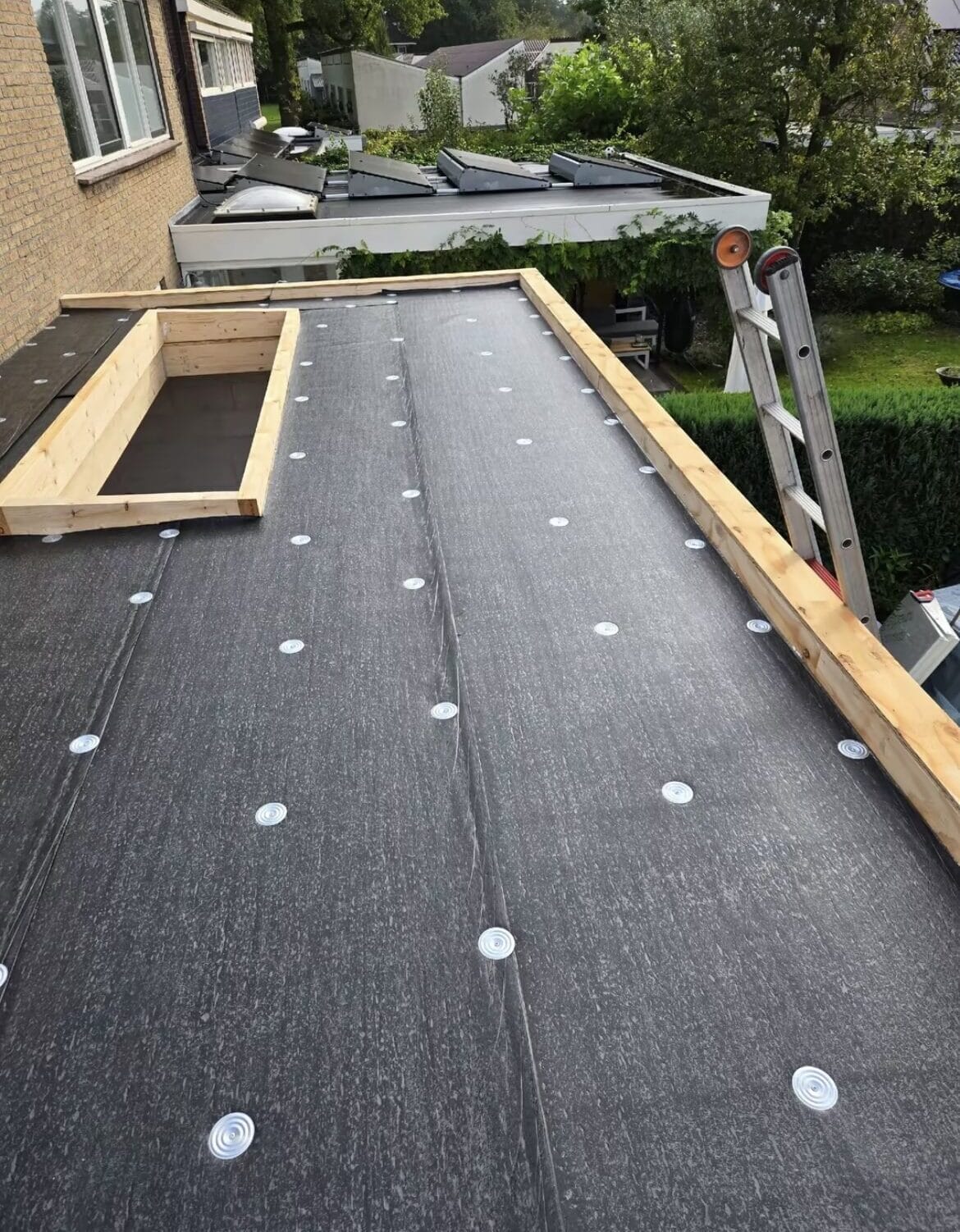 Een plat dak in aanbouw met isolatieplaten vastgezet met bevestigingsmiddelen, houten spanten aan twee kanten en een ladder die tegen de dakgoot leunt.