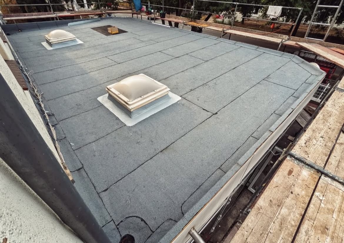 Een plat dak bedekt met asfaltvilt, met twee dakramen en omgeven door steigers en relingen.