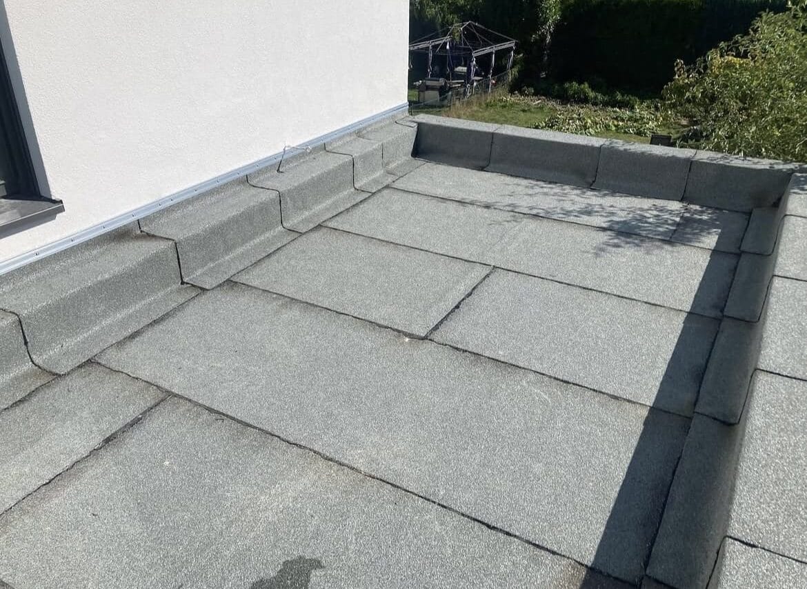 Plat dak bedekt met grijze dakbedekking, begrensd door een witte buitenmuur aan de linkerkant en omgeven door groen op de achtergrond.