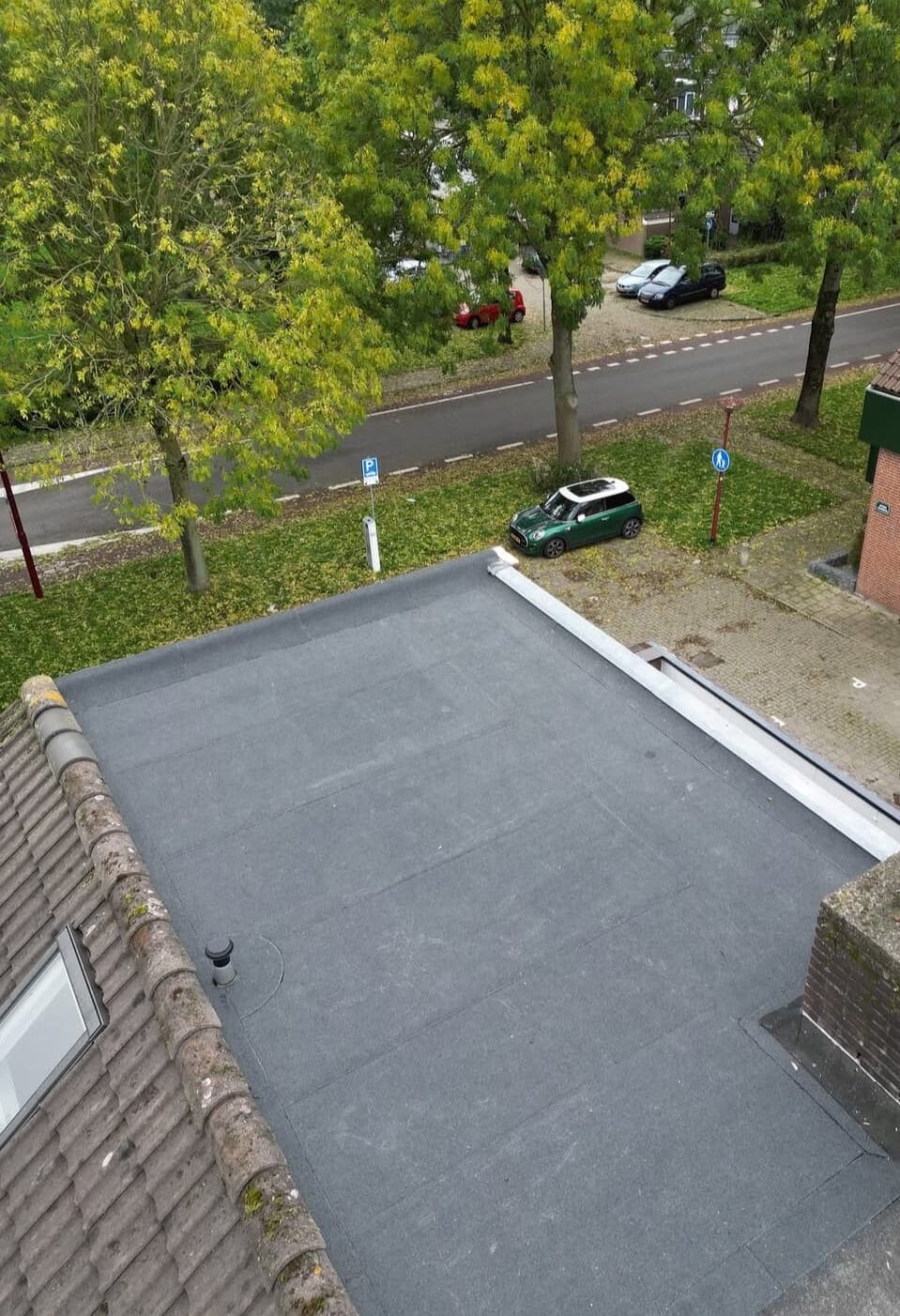 Zicht op een plat dak met grijze dakbedekking, grenzend aan een pannendak. Bomen, een weg, geparkeerde auto's en een stoep zijn zichtbaar op de achtergrond.