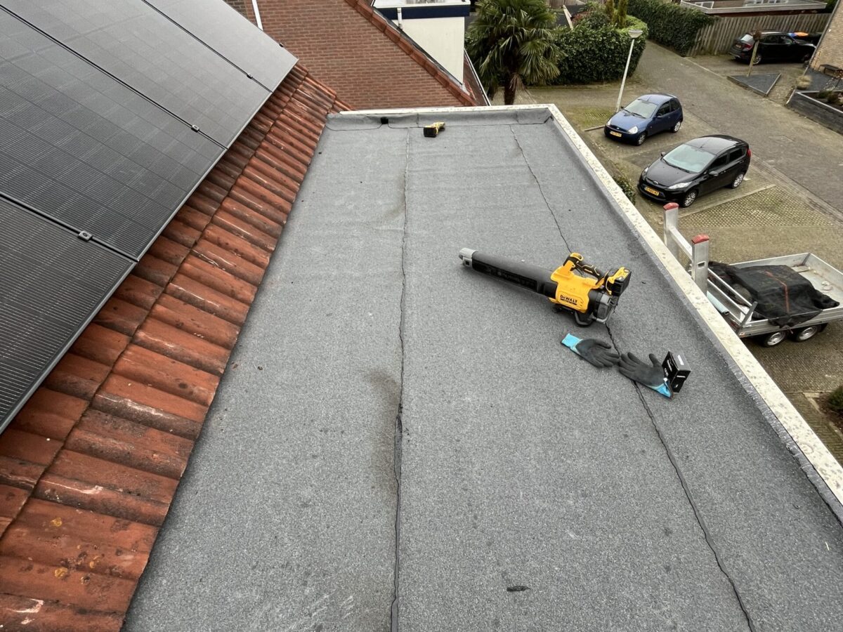 Plat dak met grijze bedekking, een bladblazer en dakgereedschap. Huizen met zonnepanelen zijn zichtbaar, naast geparkeerde auto’s en bomen op de voorstedelijke achtergrond. Plat dak met grijze bedekking, een bladblazer en dakgereedschap. Huizen met zonnepanelen zijn zichtbaar, naast geparkeerde auto's en bomen op de voorstedelijke achtergrond.