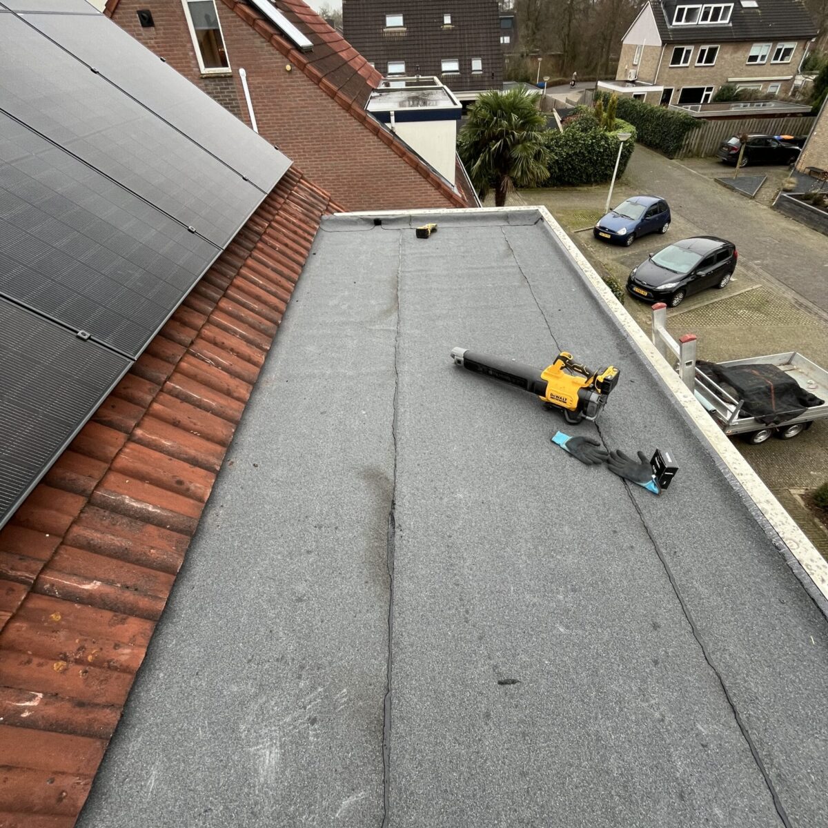 Plat dak met grijze bedekking, een bladblazer en dakgereedschap. Huizen met zonnepanelen zijn zichtbaar, naast geparkeerde auto’s en bomen op de voorstedelijke achtergrond. Plat dak met grijze bedekking, een bladblazer en dakgereedschap. Huizen met zonnepanelen zijn zichtbaar, naast geparkeerde auto's en bomen op de voorstedelijke achtergrond.