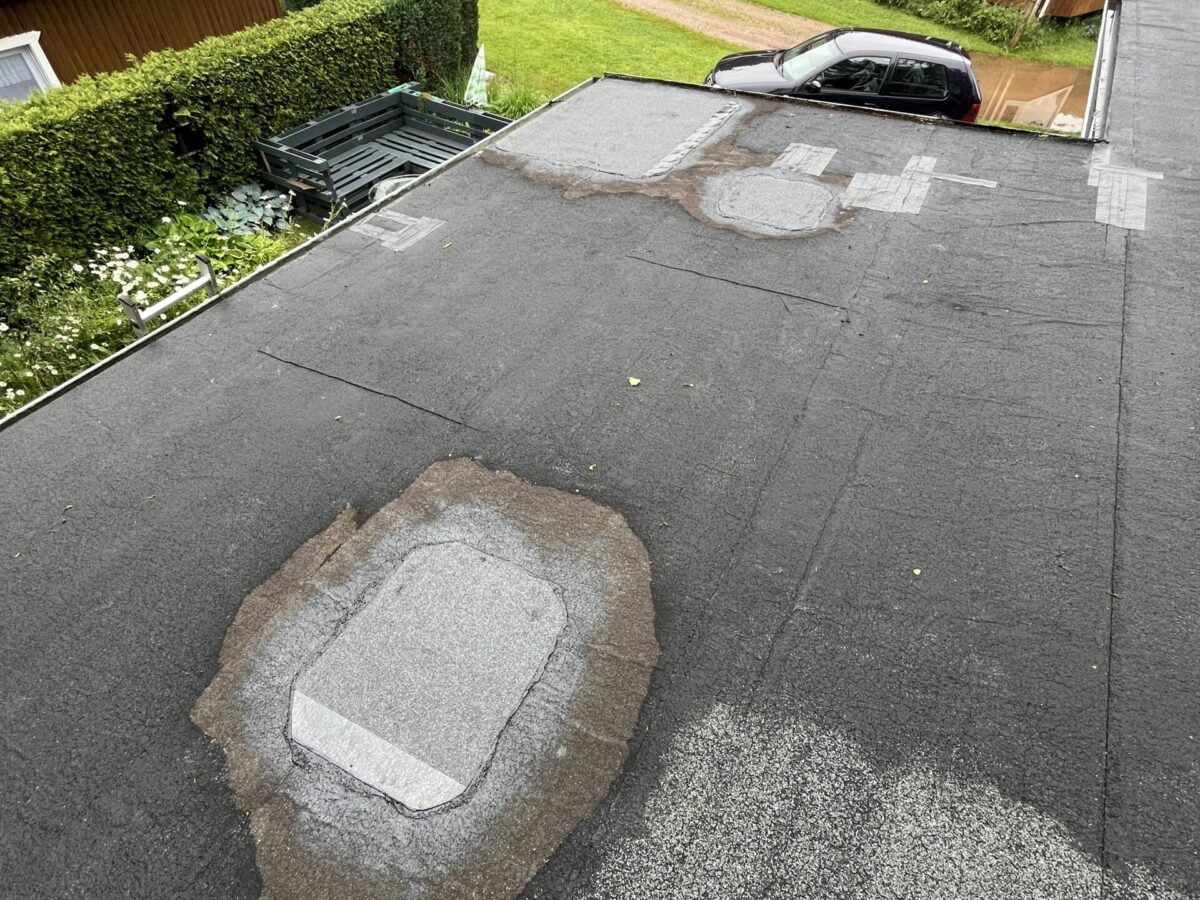 Uitzicht op een plat dak met reparaties, omgeven door groen en houten gebouwen. Een geparkeerde auto is zichtbaar bij de constructie. Uitzicht op een plat dak met reparaties, omgeven door groen en houten gebouwen. Een geparkeerde auto is zichtbaar bij de constructie.