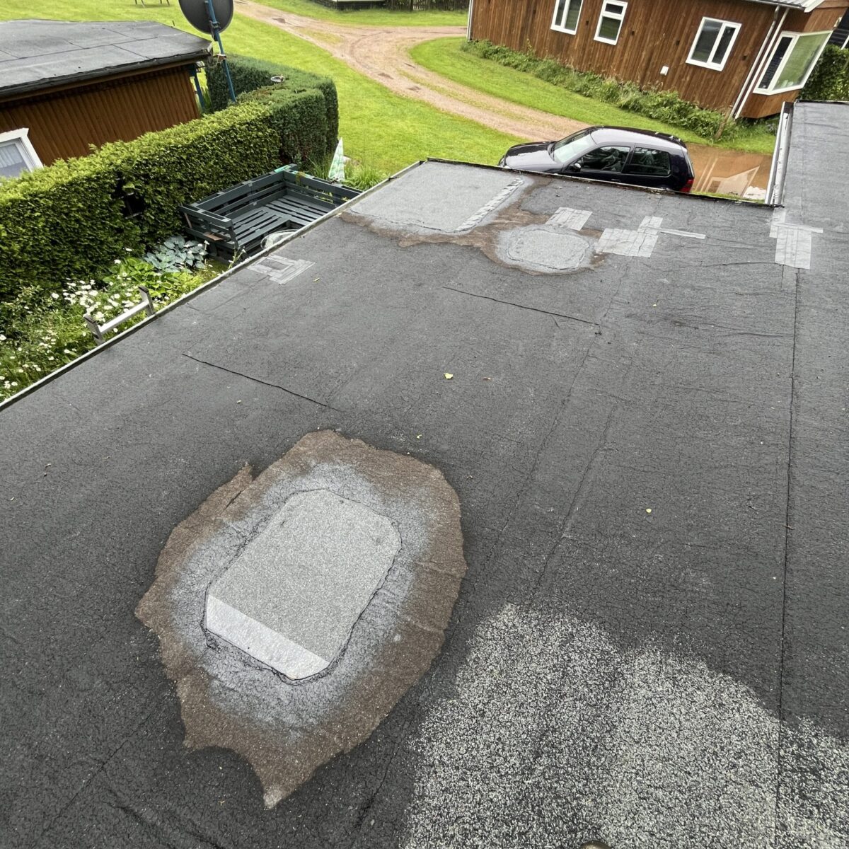 Uitzicht op een plat dak met reparaties, omgeven door groen en houten gebouwen. Een geparkeerde auto is zichtbaar bij de constructie. Uitzicht op een plat dak met reparaties, omgeven door groen en houten gebouwen. Een geparkeerde auto is zichtbaar bij de constructie.