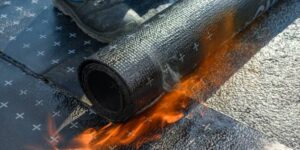 Een rol dakisolatie wordt in brand gestoken op een plat dak, met zichtbare vlammen eronder. Er staat een persoon met een laars in de buurt.