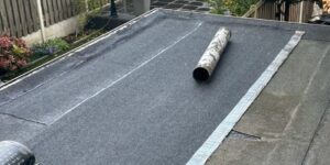 Plat dak met een gedeeltelijk afgerolde rol dakbedekking, op de achtergrond zijn de tuin en het hekwerk zichtbaar.