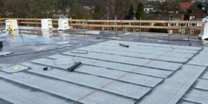 Dak met een gedeeltelijk geïnstalleerd waterdicht membraan, verspreid gereedschap en uitzicht op een bewolkte lucht en verre gebouwen.