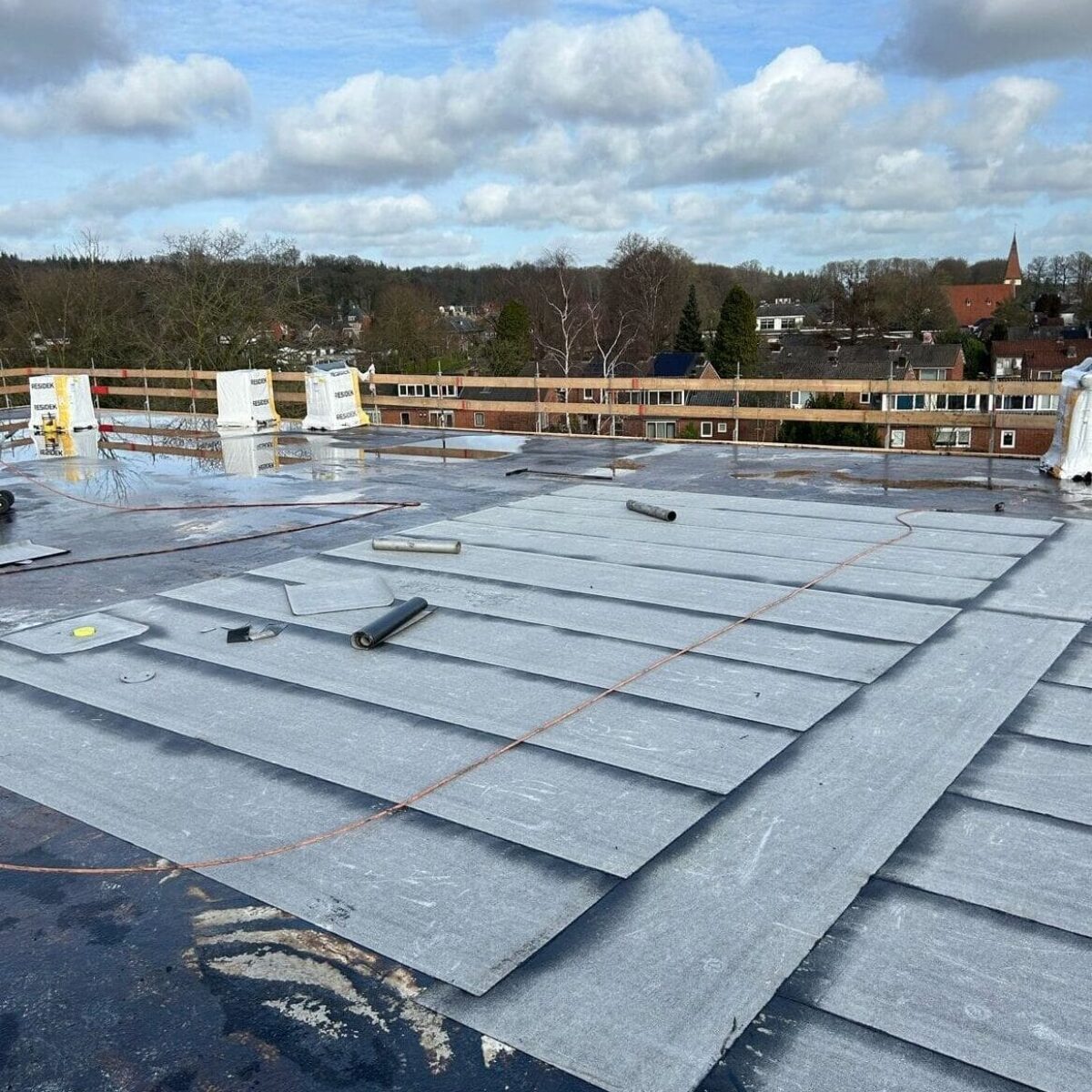 Dak met een gedeeltelijk geïnstalleerd waterdicht membraan, verspreid gereedschap en uitzicht op een bewolkte lucht en verre gebouwen. Dak met een gedeeltelijk geïnstalleerd waterdicht membraan, verspreid gereedschap en uitzicht op een bewolkte lucht en verre gebouwen.
