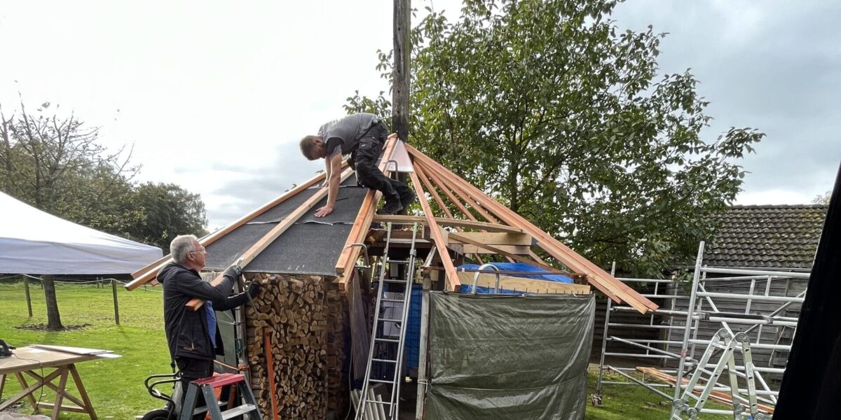 Twee mensen werken nauwgezet aan de constructie van een kleine, ronde houten constructie. Eén persoon staat op een ladder, terwijl de ander balanceert op het dakframe, waarvan een deel onder een zeil ligt. Het is bijna alsof ze een geïmproviseerde dakinspectie uitvoeren met steigers in de buurt ter ondersteuning. Twee mensen werken nauwgezet aan de constructie van een kleine, ronde houten constructie. Eén persoon staat op een ladder, terwijl de ander balanceert op het dakframe, waarvan een deel onder een zeil ligt. Het is bijna alsof ze een geïmproviseerde dakinspectie uitvoeren met steigers in de buurt ter ondersteuning.